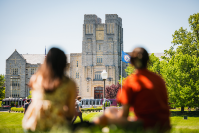 Two people sit on the Virginia Tech Drillfield with their backs to the camera, facing Burruss Hall. VT buses and the Virginia state flag are visible infront of the building. A cloudless, light blue sky is silhouetted behind and flanked by green trees.