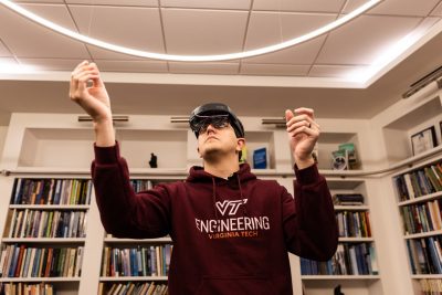 A man in a maroon VT Engineering hooded sweatshirt wears a headset with glasses while reaching toward the ceiling.