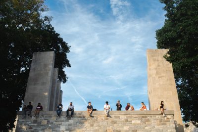 Twelve people sit evenly spaced across the wall above the War Memorial Chapel on the Virginia Tech Drillfield. The Pylons are visible behind them against a blue sky with high wispy clouds, flanked by green trees.