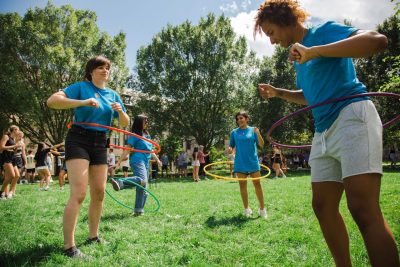Four women in blue shirts play with hula hoops on a green lawn, surrounded by green trees on a sunny day.