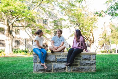 Three students talk while sitting on a Hokie Stone wall on the Drillfield at Virginia Tech.
