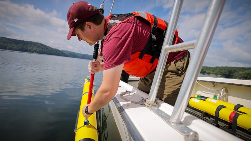 A man wearing a maroon hat and an orange life jacket holds a yellow tube over the side of a boat while on a lake.