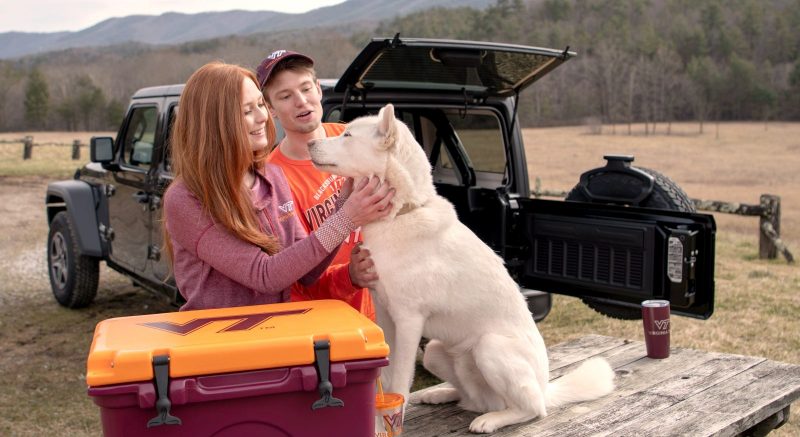 Two students in Virginia Tech apparel with a white dog and Virginia Tech cooler.